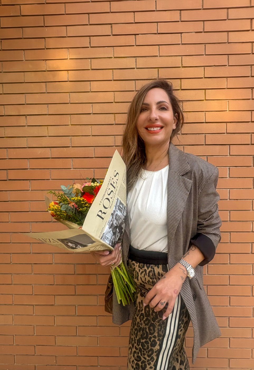 Η Σοφία χαμογελά, με λουλούδια, μπροστά σε τοίχο από τούβλα