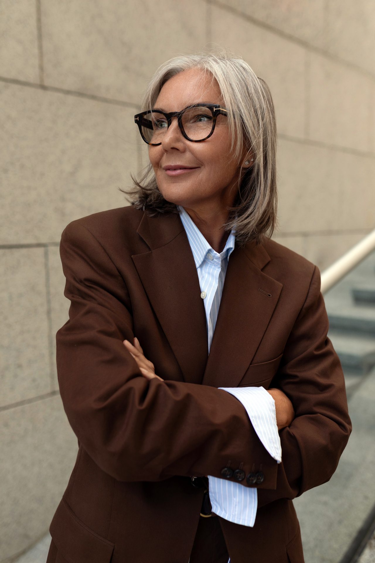 A woman with grey hair, glasses and a brown blazer